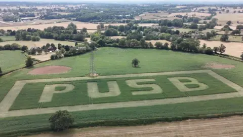 Aerial view of countryside with trees and hedges and green and yellow fields. The letters P U S B are cut into the nearest field.