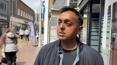 LDRS A man wearing a grey top and jacket. He is stood in a high street looking away from the camera.