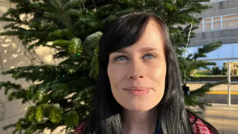 John Devine/BBC Gemma, a woman, standing in front of the Christmas tree, smiling for the camera.