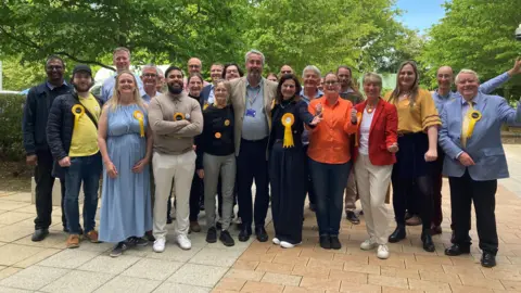 Emma Howgego/BBC A large group of people wearing Liberal Democrat rosettes smile and cheer. 