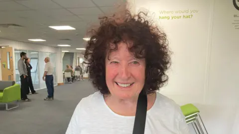 A photo of a woman with dark frizzy hair. She is wearing a white T-shirt and is standing in front of a wall. To the side a number of people are standing and chatting.