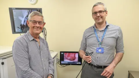 Peter Russel stands next to ENT consultant Mr Kostas Marinakis. Peter is wearing a white and blue striped shirt and has grey hair and glasses. Kostas is also wearing a white and blue striped shirt and has grey hair and glasses. Kostas is holding a scoping device in his hand and in the middle of the two men is a screen which appears to show the inside of someone's throat.