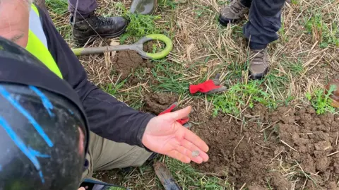 Worcestershire County Council A metal detectorist holds a coin in a palm of their hand. Below them is a muddy hole in the ground. Around them can be seen the feet of other people at the scene. 