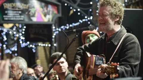 Getty Images A man with curly, reddish-grey hair is playing guitar and singing. He has a semi-acoustic guitar in his hands, his eyes are closed mid-singing, there is a microphone close to his mouth and in the background are people, small lights and signs for a building. The scene appears to be on a street at night time. 