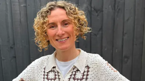 Person with short, tightly curled hair standing in front of a dark wooden fence, wearing a cream crocheted short-sleeve shirt with brown square patterns over a light-colored top.