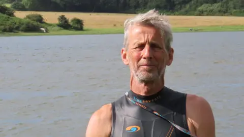 Ric Cooper of The Cleddau Project ready to go for a swim in the Western Cleddau river.  He's standing in front of the river wearing a black, sleeveless wetsuit. He has grey short hair and grey stubble. 
