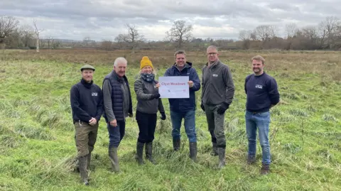 Ben Morgan-Brown, Councillor Geoff Jung, Councillor Paula Fernley, Councillor Henry Massey, Paul Osborne and James Chubb pictured standing in a large grassy field. They are wearing warm clothing and wellington boots while holding a large sheet of paper that reads Clyst Meadows.