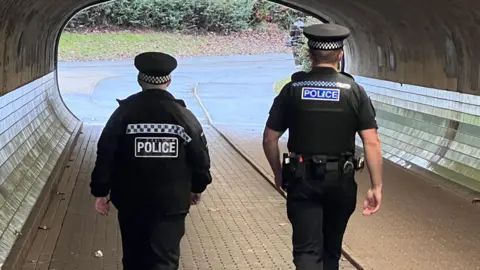 Simon Dedman/BBC Two police officers walking through an underpass and away from the camera; they have their backs to us. Both of them are in uniform. The officer on the left is slightly shorter and is wearing a dark police officer's uniform, with a long-sleeved jacket and a peaked cap. The officer on the right is taller, is also wearing a uniform, though he is wearing a t-shirt and his bare lower arms can be seen. The pair are walking on paving stones towards the bright tunnel exit. The walls are curved, with the lower section covered with shiny tiles. 