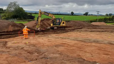 Oxford Cotswold Archaeology An archaeology site which has brown-reddish coloured earth. There are little blue flags and small parts which have been dug up. Two people wearing orange fluorescent overalls work on the site.