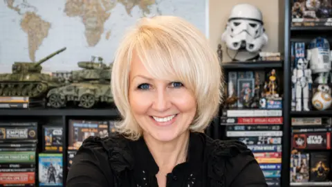 Dennie Smith Dennie Smith with a blonde looks into the camera and smiles. Behind her are models of tanks and Star Wars merchandise.