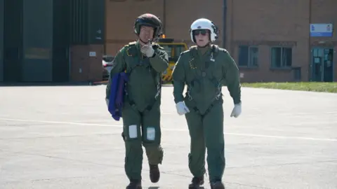 An instructor and student in green jumpsuits and flight helmets walking towards an unseen aircraft. 