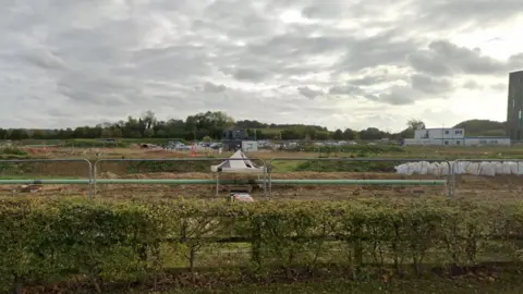 Google A piece of land fenced off by developers on Dame Mary Archer Way. In the foreground is a hedge and fence and beyond that metal fencing, scrubby land, rows of aggregate bags on the right. Behind them are portable cabins and on the horizon trees and sky. 