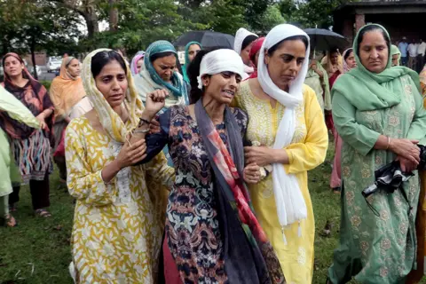 Reuters A group of women dressed in traditional clothing and headscarves, standing outdoors with trees and a building in the background. The woman in the centre - Vihaan's mother - is wailing. 