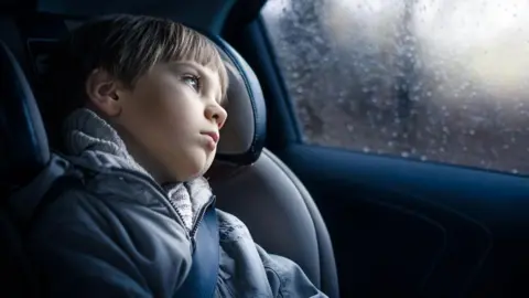 A child sits secured in a car seat in the back of a vehicle, wearing a grey coat and seatbelt, looking out through a rain-speckled side window. The interior of the car is dimly lit, with soft daylight filtering through the window and blurred scenery visible outside.