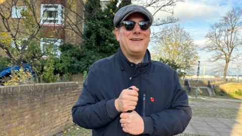 Simon Kennedy standing on a pavement, wearing flat tweed cap, dark glasses and a navy jacket, grasping a cane in both hands. He is smiling at the camera.