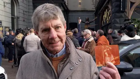 Ciaran Laverty who has short grey/brown hair and is wearing a light brown coat with a brown jumper and blue shirt underneath. He is holding up a red-stained photo of Rory Gallagher performing on stage with his guitar. Mr Laverty is standing in front of the Ulster Hall with the statue behind him. 