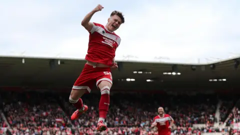 A footballer in the red shirt and shorts of Middlesbrough leaps into the air punching his fist. A teammate is running towards him smiling. In the background is a full stand of cheering fans.