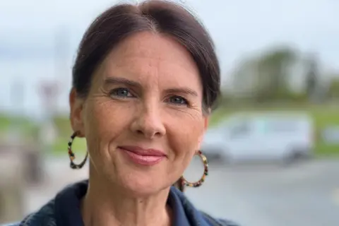 Trudy Harrison is looking straight ahead, wearing a denim jacket. She has dark hair, tied back, and is wearing medium sized gold hoop earrings.