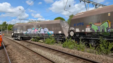 Two large freight wagons with their wheels off the tracks and overhead power lines above
