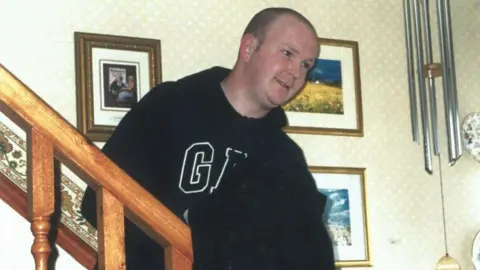 Pacemaker Glenn Quinn, with short grey hair, wearing a black GAP hoodie. He is standing on a stairs in a home. There are framed paintings and photos on the wall behind him. 