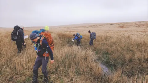 Mount Kelly College Four people scattered in long grass all looking down. They are wearing hats and rucksacks.