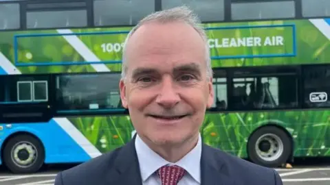 Chris Conway standing in front of a Translink bus in a suit with a red tie