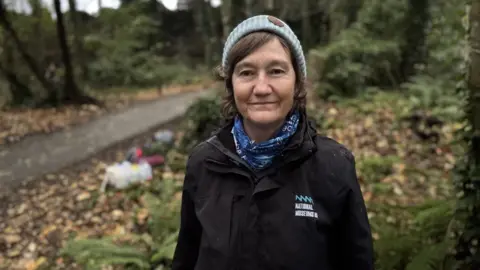Helen James, a woman with dark hair, dark jacket, light blue hat and darker blue scarf. She is standing in a forest area.