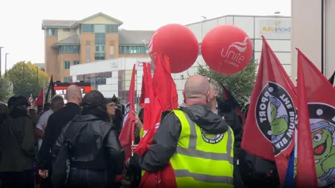 The backs of a crowd who are holding red flags and balloons