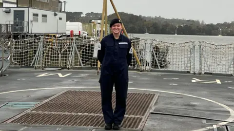 A woman smiling whilst standing onboard the HMS Richmond. The ocean and hills can be seen in the background. Grey skies.
