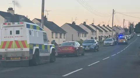A police land rover parked at the side of the road beside a row of houses. A number of other cars are parked further along the road, one of them a police car.