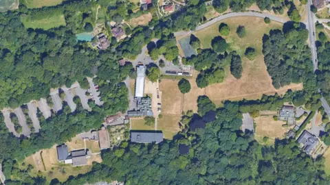 An aerial image of the site which shows trees and green spaces as well as several buildings and a large carp park. There are tennis courts visible.