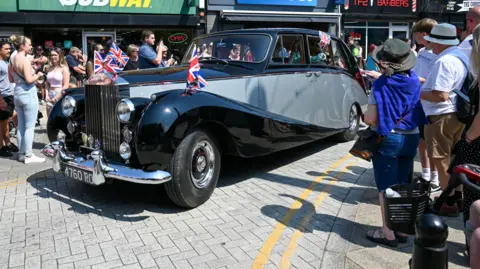 Shropshire Festivals A vintage car with grey and black markings and union flags in the front. A crowd of people are surrounding the vehicle. 