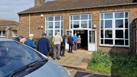 Anthony Kimber Residents of Rye, East Sussex, queue at their local vaccination centre with the double doors to the building open on a sunny day.