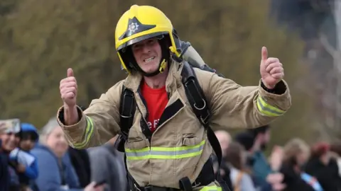 Chris Dabell Chris Dabell is running with his thumbs up whilst wearing full firefighters uniform - including a yellow helmet.