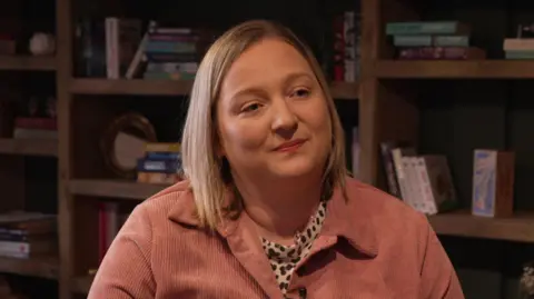The image shows a woman seated indoors in front of a large wooden shelving unit. The shelves are arranged in a grid pattern and contain a mixture of books, decorative ornaments, small vases, framed pictures, and plants. The person is wearing a light, textured button‑up jacket in a muted pink shade over a patterned top with black and white markings. A small clip‑on microphone is attached to the jacket