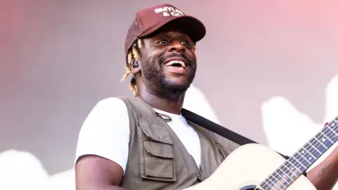 Getty Images Myles Smith performing during 2024 Austin City Limits Music Festival at Zilker Park in October 2024 in Austin, Texas. He's holding a guitar and smiling, whilst wearing a brown cap, and a white t-shirt. 