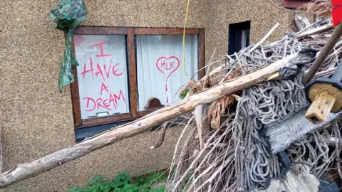 Denis Carbonaro "I have a dream" written in red ink on a window next to a heart with MLK in the middle of it. In front of the window there is a pile of wood and ropes