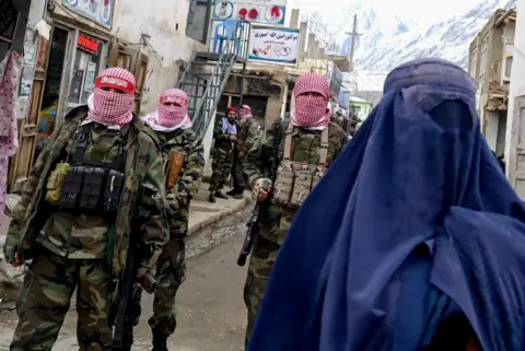 AFP Taliban security personnel stand guard as an Afghan burqa-clad woman (R) walks along a street at a market in the Baharak district of Badakhshan province on February 26, 2024
