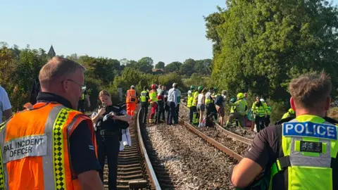 Rob Bradley Emergency service personnel, rail staff and passengers stand on a railway line near Lewes in East Sussex following the derailment. Police officers and rail incident officers are visible in the foreground while groups of people gather further along the track during the response to the incident.