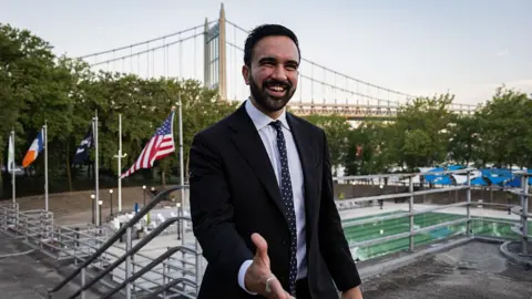 Getty Images Zohran Mamdani reaches out his hand. He is wearing a suit. A bridge can be seen behind him, along with flags