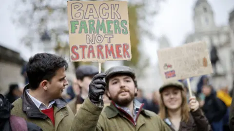 EPA Three farmers, hold signs at the protest. The central sign, held by a man wearing a tweed coat and a flat cap reads 'back farmers not Starmer'