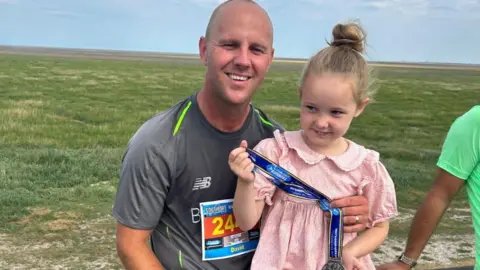 Family handout David and his daughter Elsie in 2023 after David had completed the Southport 10k. Elsie is wearing a pink dress with her hair in a bun and holding David's medal while David wears a grey running top and smiles at the camera.