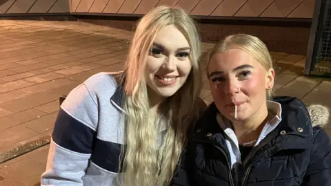 18-year-old friends Lara Smith and Imogen Hanna sitting together on a bench at Southwater. One of the teenagers has a lollipop in their mouth which was handed to her by the street pastors.