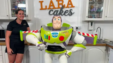 A woman standing next to large Buzz Lightyear cake. The woman has brown hair tied back and the cake is white and green with three small buttons which are blue, green and red and a large red button. The cake also has wings which are striped red and white. 