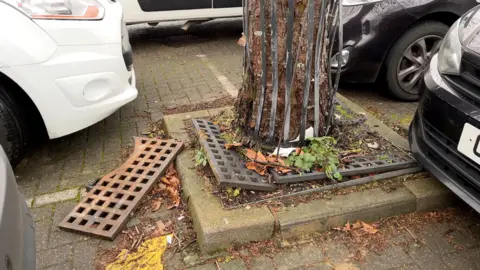 BBC The base of tree that has broken through its ironwork surrounds, causing a trip hazard. One piece of iron grid lies next to the tree, straddling car park spaces.