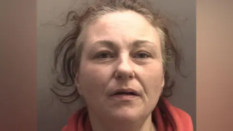 A woman in her 40s, with unkempt brown hair and wearing a red fleece, looks into the camera with a slight smile