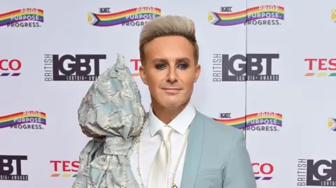 Getty Images H from Steps wearing heavy eye make-up while smiling on the red carpet. He has a light blue suit with a large lacey bow on the left shoulder