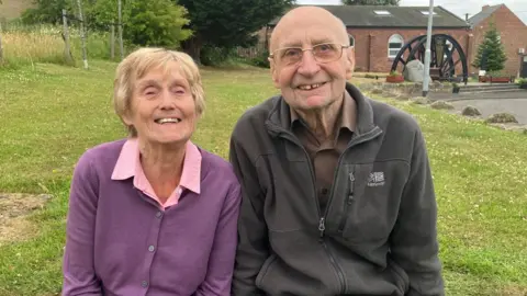 Head and shoulder shot of two elderly people. The woman, on the right is wearing a purple cardigan over a pink blouse and the man, who is balding, is wearing spectacles and a brown zipped-up fleece over a brown shirt. Both are smiling, and behind his right shoulder can be seen brick buildings with a set of pit wheels embedded in the ground in front.