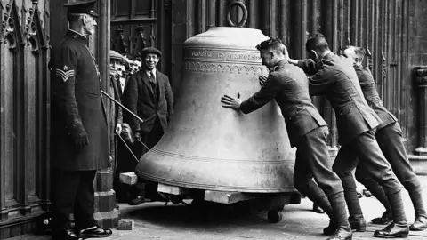 Getty Images Uniformed individuals push a massive metal bell on wheels outside a large stone building with columns. Several onlookers stand nearby, watching as the bell is carefully moved into position. The black-and-white scene suggests a historical moment involving the transport or installation of the b
