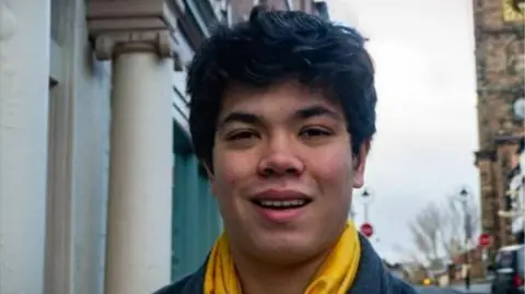 A man with black hair, a yellow scarf and dark coat standing in a town street with a church behind him.